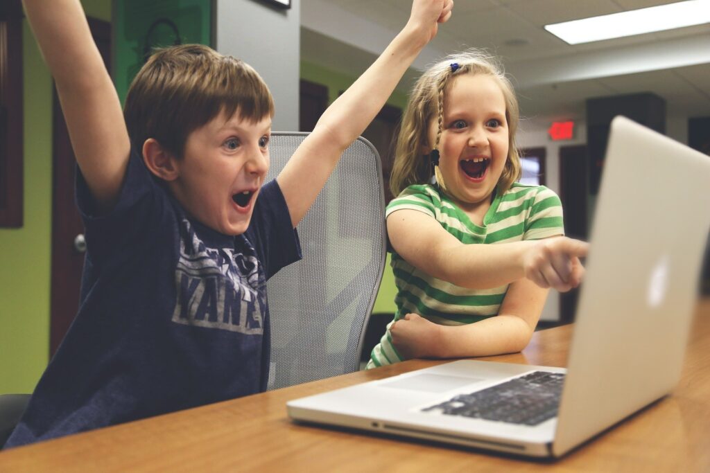 Chłopiec i dziewczynka siedzą przed laptopem. Chłopiec z radości unosi ręce do góry, dziewczynka z uśmiechem wskazuje palcem na monitor