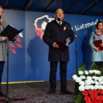 Burmistrz Słupcy, Wójt Gminy Strzałkowo i dyrektor GOKiS stoją na scenie podczas obchodów Narodowego Święta Niepodległości w Łężcu. Przed nimi biało-czerwone kwiaty ułożone w kształcie flagi Polski