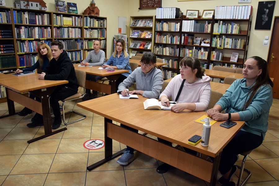Siedem osób siedzi za stołami w czytelni biblioteki. Wokół regały z książkami