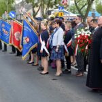 Kilkuosobowa grupa uczestników uroczystości stoi na ulicy przed pomnikiem Powstańców Wielkopolskich