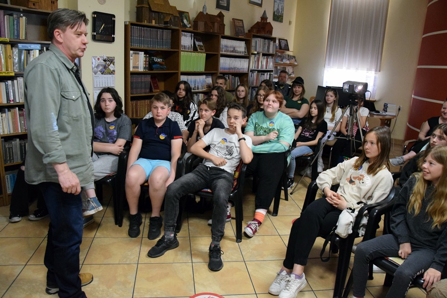 Grupa młodzieży siedzi na krzesłach w czytelni biblioteki słucha prowadzącego spotkanie, który stoi przed nimi