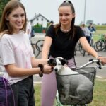 Dwie uśmiechnięte młode dziewczyny na rowerach. U jednej w rowerowym koszyku siedzi mały, biało-czarny pies