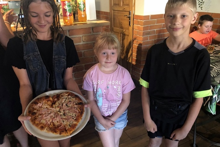 Dwie dziewczynki i chłopiec w pizzeri. Jedna z dziewczynek prezentuje pizzę w kształcie serca