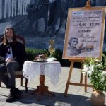 Pracownik biblioteki wcielający się w postać Adama Mickiewicza siedzi przy okrągłym stoliku. Obok na sztaludze plakat akcji Narodowe Czytanie