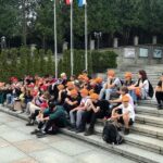 Uczestnicy wycieczki siedzą na betonowych schodach przed sanktuarium na Krzeptówkach słuchając przewodnika
