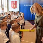 Wicedyrektor szkoły podstawowej w Strzałkowie pasuje pierwszoklasistów na uczniów dotykając ramienia dziecka wielką, kolorową kredką