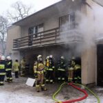Strażacy w maskach tlenowych podczas ćwiczeń przed domem, z którego wydobywa się dym