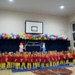 Dzieci z wszystkich grup razem z opiekunkami stoją trzymając się za ręce w sali gimnastycznej. Za nimi dekoracja z balonów z napisem "Pasowanie na przedszkolaka"