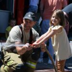 Strażak pomaga dziewczynce utrzymać wąż strażacki, z którego leje się woda