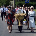 Kilkadziesiąt osób idzie ulicą. W pierwszym rzędzie mężczyzna niesie dożynkowy wieniec