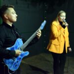 Młody mężczyzna na scenie gra na gitarze, a kobieta śpiewa do mikrofonu