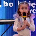 Dziewczynka śpiewa do mikrofonu. Za nią widać fragment pianina elektrycznego