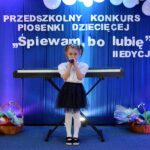 Dziewczynka śpiewa do mikrofonu na tle napisu z nazwa konkursu