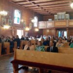 Kilkadziesiąt osób siedzi w ławkach w drewnianym kościele słuchając koncertu