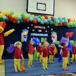 Grupa dzieci w żółto-czerwonych strojach oraz maskotka Kubusia Puchatka tańczą z kolorowymi chustkami