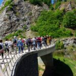 Grupa uczestników wycieczki przechodzi przez betonowy most w dawnym kamieniołomie. Przed nimi wznosi się wielka skała