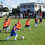 Akcja podczas meczu. Młody zawodnik w niebieskiej kamizelce biegnie z piłką przy nodze, a czterech zawodników w pomarańczowych kamizelkach biegnie w jego stronę