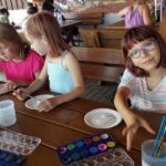 Trzy dziewczynki siedzą przy stole i wykonują prace plastyczne z użyciem farb