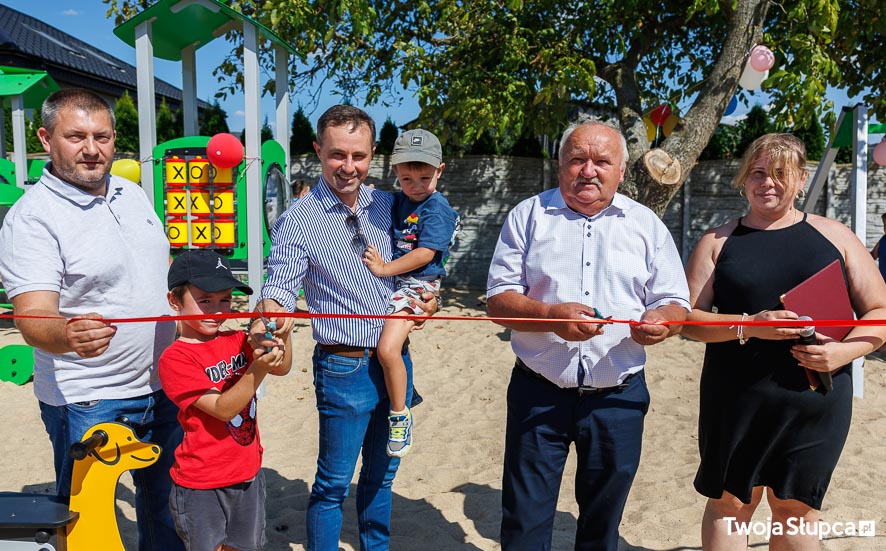 Wójt Gminy, sołtys, radny i przedstawicielka mieszkańców przecinają wstęgę na nowym placu zabaw. Wójt trzyma na ręce chłopca. Obok niego drugi chłopiec pomaga mu w przecięciu wstęgi