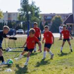 Akcja podczas meczu młodych piłkarzy. Zawodnik w czerwonej koszulce próbuje odebrać piłkę bramkarzowi drużyny przeciwnej