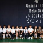 Pierwszoklasiści siedzą na ławce na tle napisu Gminna Inauguracja Roku Szkolnego 2024/2025