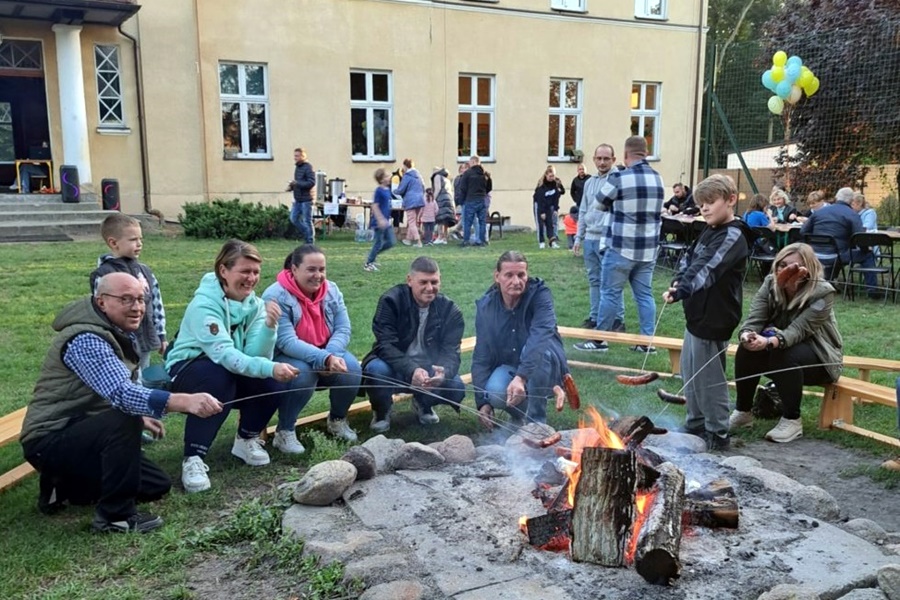 Grupa osób siedzi przy ognisku i piecze kiełbaski