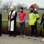 Wójt Gminy Strzałkowo, Burmistrz Miasta Słupcy i Starosta Powiatu Słupeckiego w sportowych strojach stoją w rzędzie. Obok stoi ksiądz w stule oraz dyrektor Miejskiego Ośrodka Sportu i Rekreacji w Słupcy, która mówi do mikrofonu