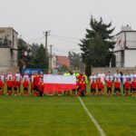 Na środku boiska stoją zawodnicy obu drużyn i sędziowie. Młodzi zawodnicy trzymają rozwiniętą biało-czerwoną flagę