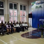 W sali gimnastycznej wójt przemawia do mównicy zwracając się w stronę stypendystów z ZSP Strzałkowo siedzących na krzesłach