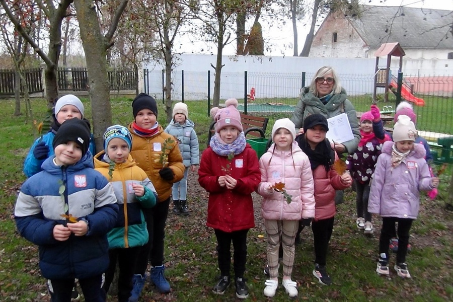 Grupa dzieci wraz z nauczycielką pozuje do wspólnego zdjęcia. W rękach trzymają zebrane liście