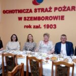 Pięć kobiet i jeden mężczyzna siedzą przy zastawionym stole. Za ich plecami na ścianie znajduje się napis Ochotnicza Straż Pożarna w Strzałkowie rok zał. 1903