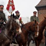 Trzech mężczyzn w powstańczych mundurach na koniach. Jeden z nich trzyma w górze sztandar