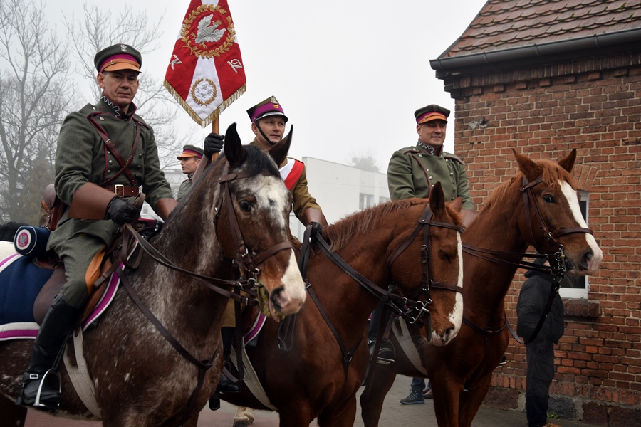 Trzech mężczyzn w powstańczych mundurach na koniach. Jeden z nich trzyma w górze sztandar