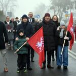 Uczestnicy uroczystości stoją na ulicy. W pierwszym rzędzie przedstawiciele samorządu gminy oraz dwójka dzieci trzymających powstańcze flagi